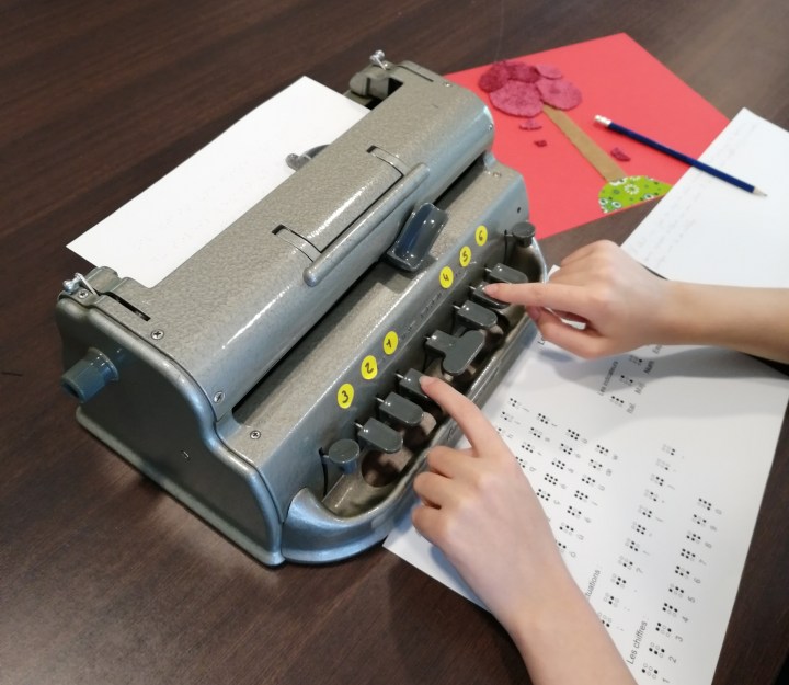 deux mains écrivent sur une machine perkins
un alphabet braille est posé sous la machine. sur le côté, une illustration en relief représentant un arbre aux feuilles tombantes