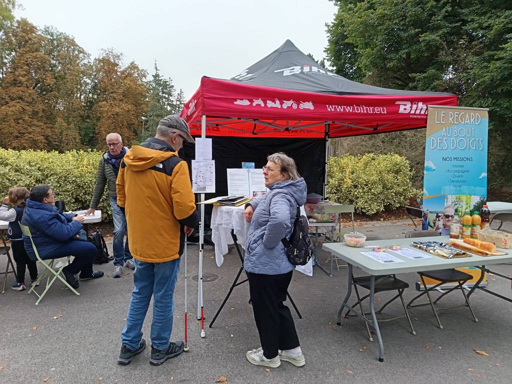 fabrice renseigne une personne devant le stand de l'association (tonnelle et tables sont disposés)
