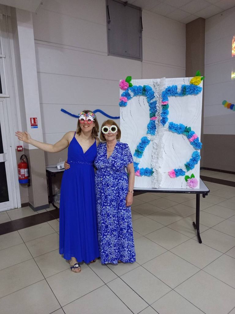 aurore et Françoise vétues d'une robe bleue et de lunettes festives devant un panneau géant écrit 25 en fleurs en papier bleues